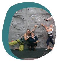 Drei Frauen stehen vor einer Kletterwand in einer Sporthalle. Zwei Frauen umarmen sich und lächeln in die Kamera, die dritte Frau lacht und schaut nach oben, wo eine weitere Person an der Kletterwand hängt.