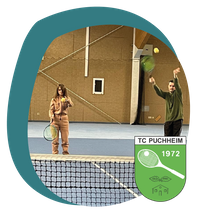 Eine blinde Frau und ein blinder Mann stehen auf einem Tennisfeld in einer Turnhalle. Sie haben Tennisschläger in der Hand und spielen über das Netz, das im Vordergrund zu sehen ist.